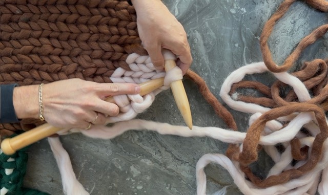 Knitting a cream chunky cardigan piece on a pair of giant 25mm birch needles, with soft roving trailing across a stone surface.