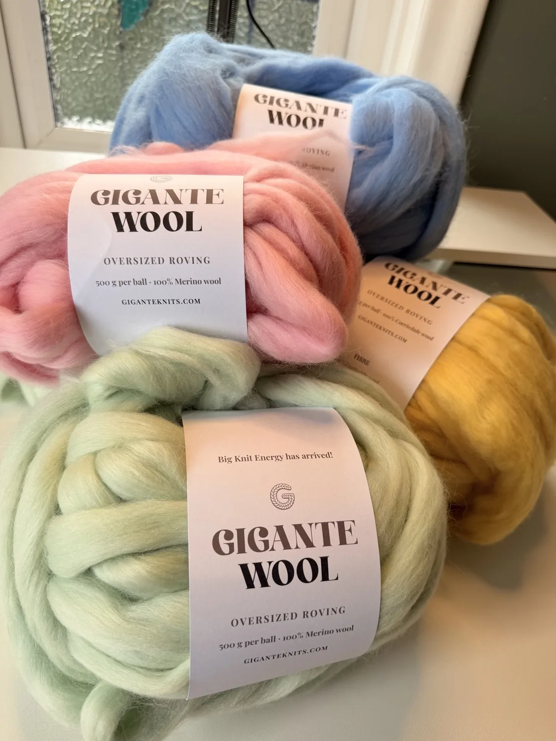 Four balls of Gigante Wool oversized roving stacked by a window: soft blue, candy pink, pale sage green and warm mustard.
