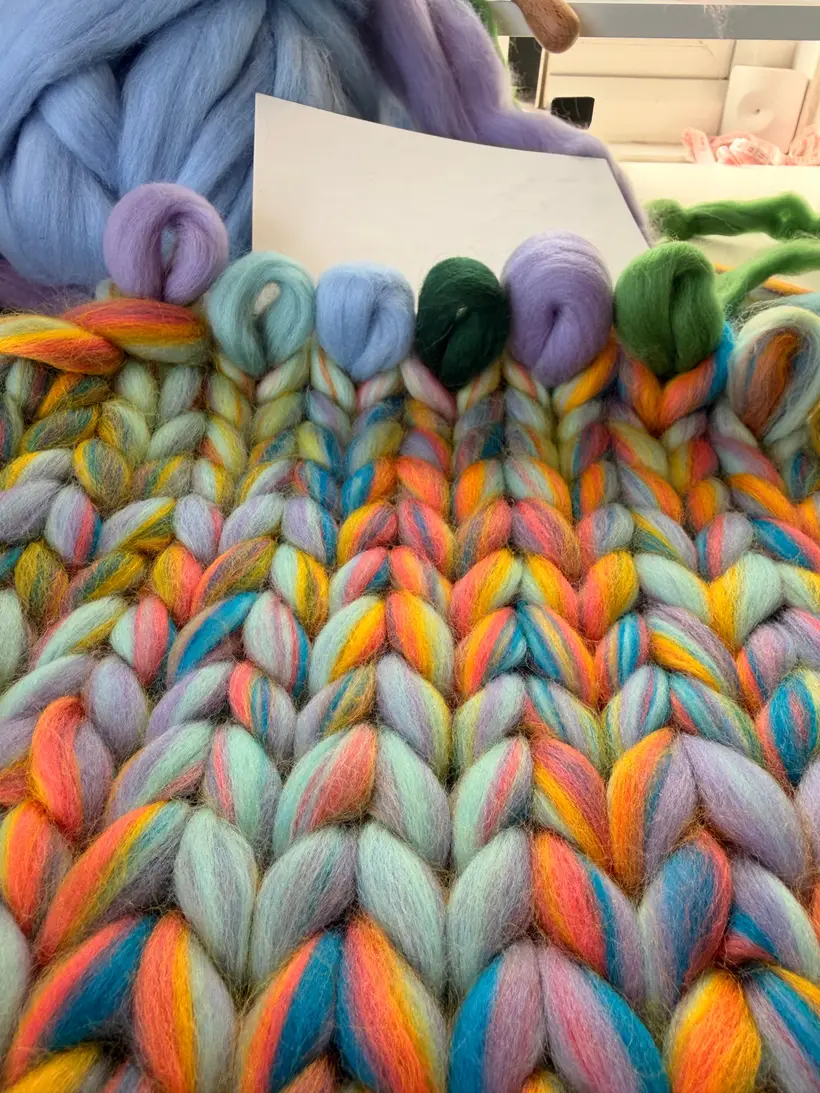 Løkker af uspundet uldtop i Lavender, mint, himmelblå, skovgrøn, lilla og bladgrøn lagt langs toppen af en Pastel Rainbow-strikkeprøve, farveprøverne til båndet.