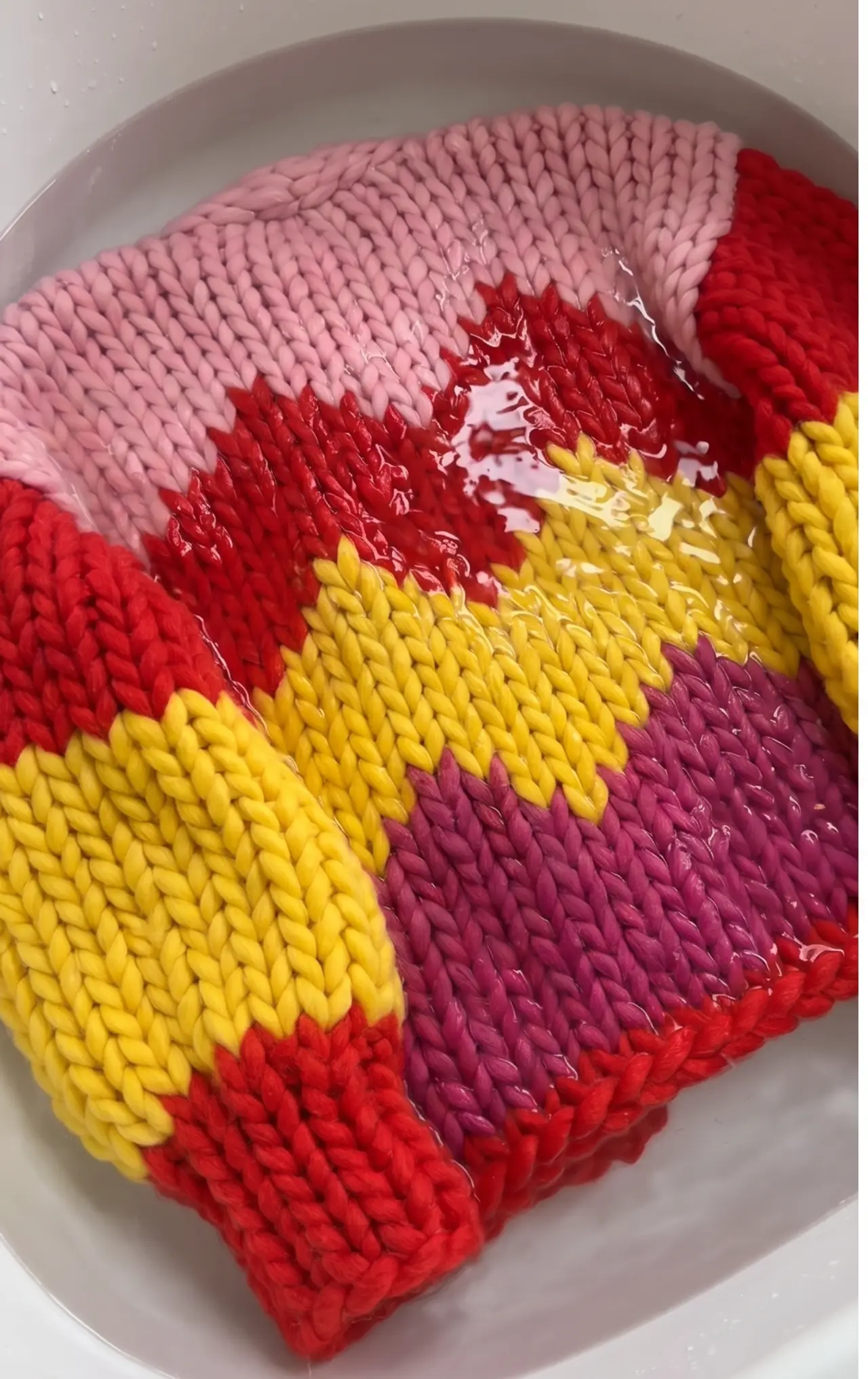 A macro close-up of chunky-knit stitches in pink, red, yellow and purple, fully soaked in warm water during blocking.