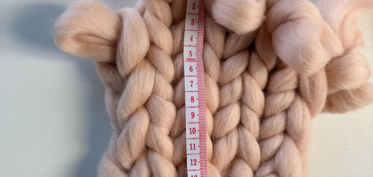 A blush-pink chunky knit gauge swatch laid flat on a white table, with a tape measure running down it reading about 16 centimetres.