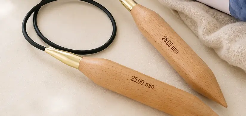 Two jumbo beech wood circular knitting needles engraved 25.00 mm, resting beside balls of Gigante Wool.