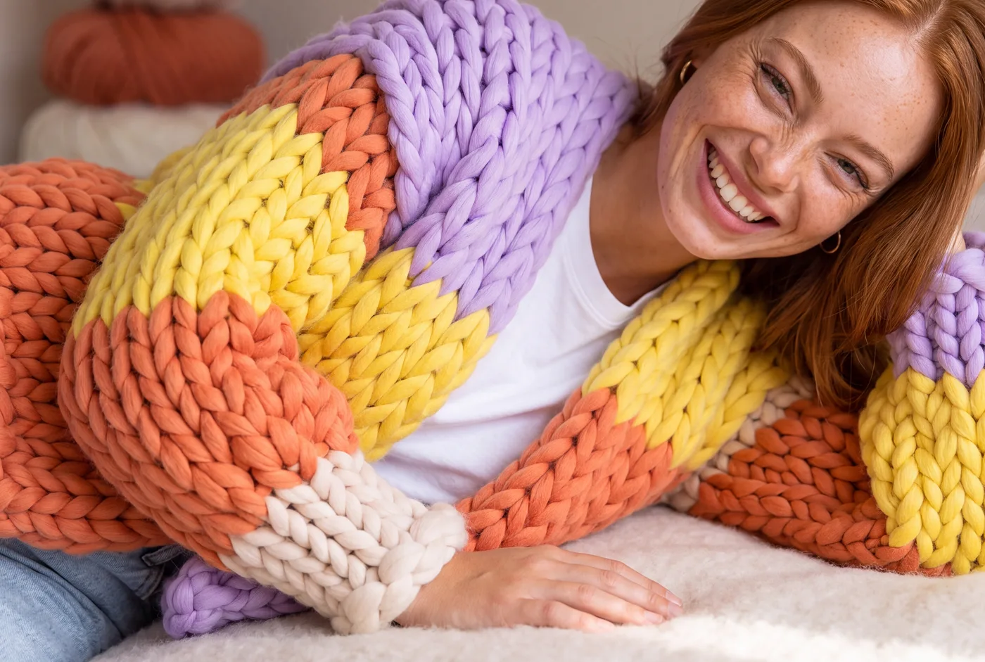 A woman lying on a bed, smiling, in an oversized chunky-knit Gigante cardigan in lilac, orange, yellow and cream