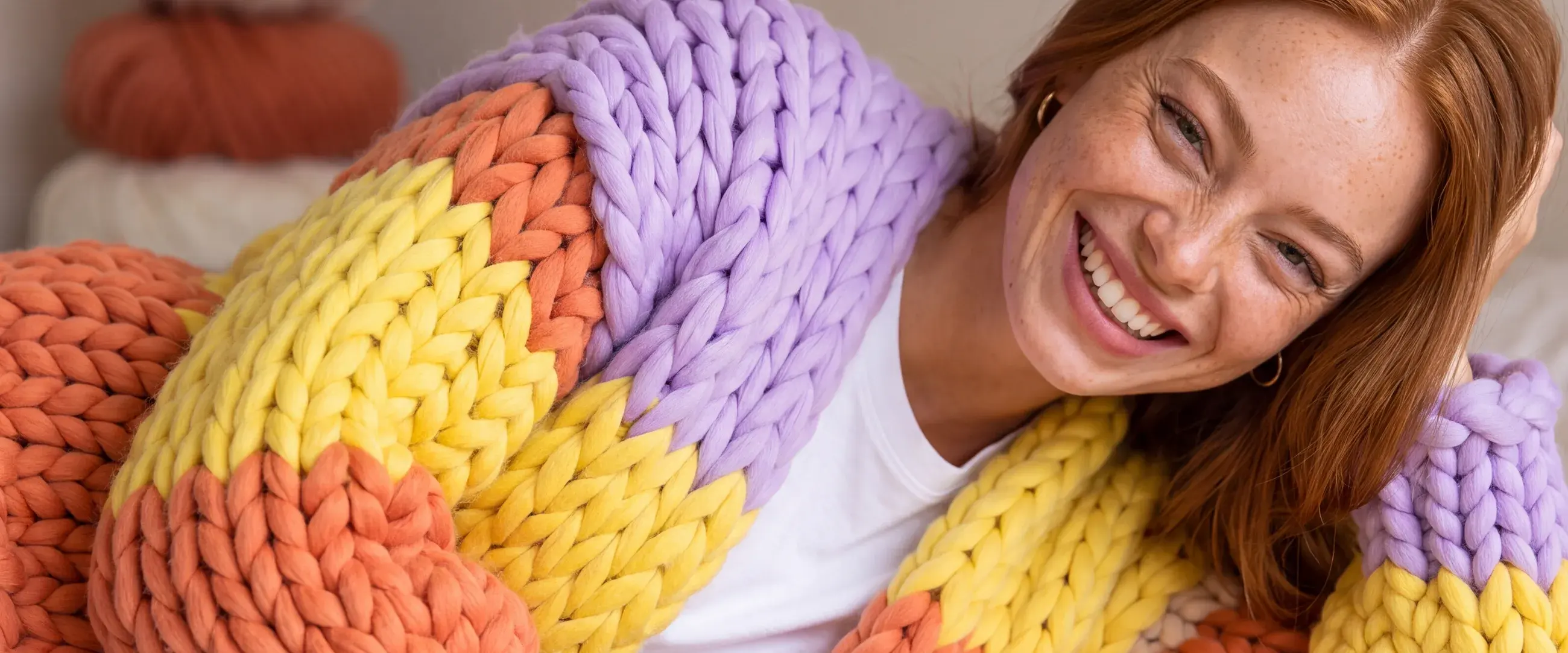 A woman lying on a bed, smiling, in an oversized chunky-knit Gigante cardigan in lilac, orange, yellow and cream