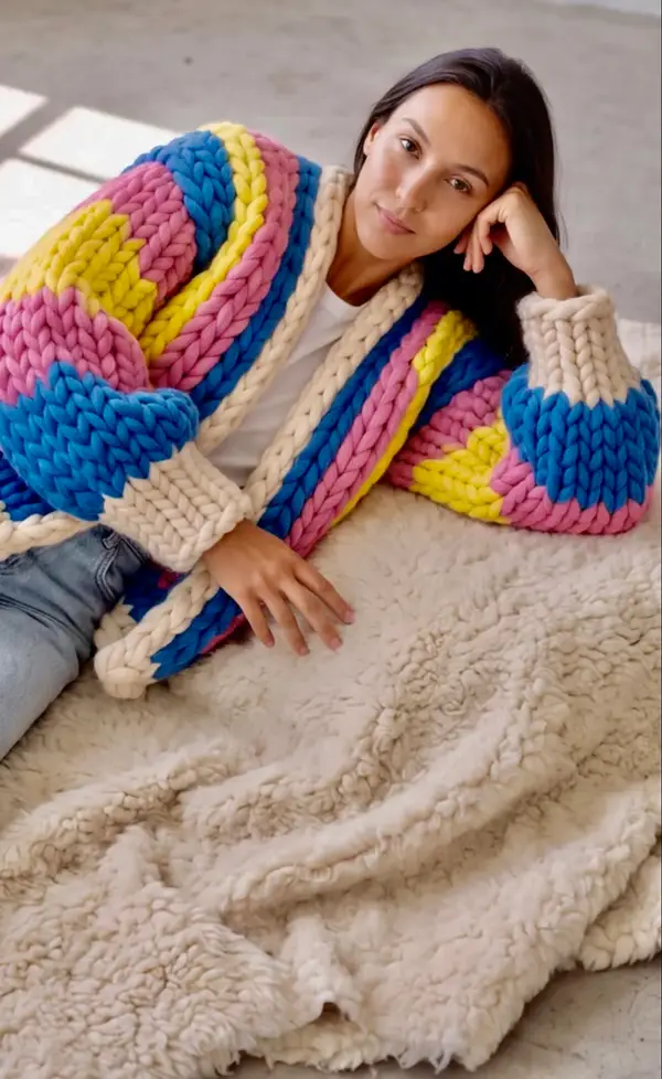 A woman lying on a sheepskin rug in the Retro Pop cardigan, propped on one elbow.